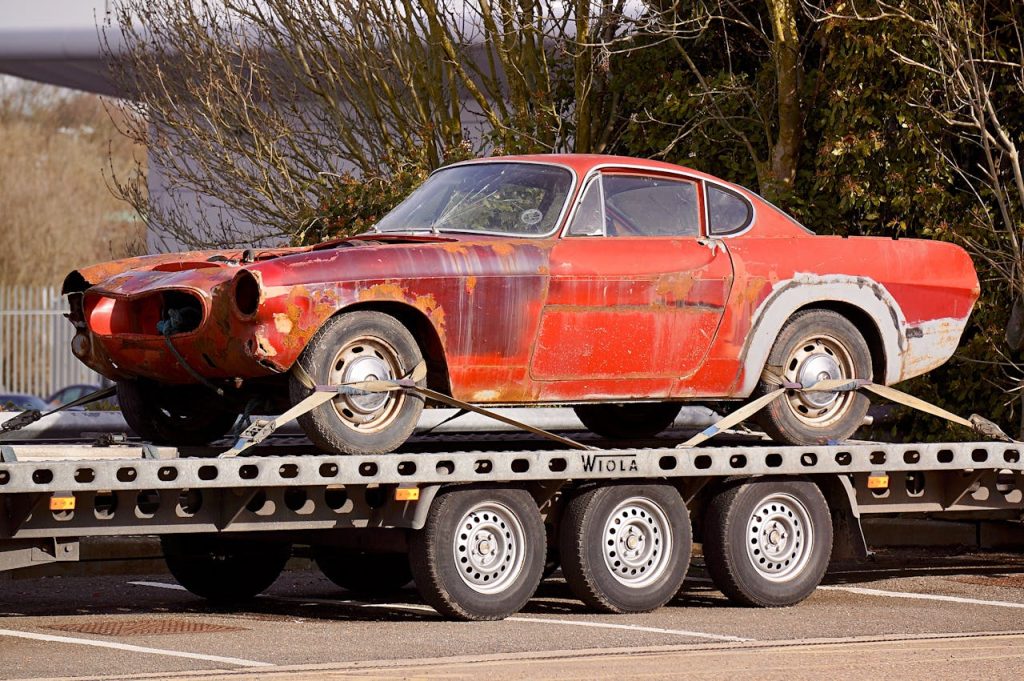 Offerings A worn-out, rusty vintage car secured on a flatbed trailer, outdoors under daylight.